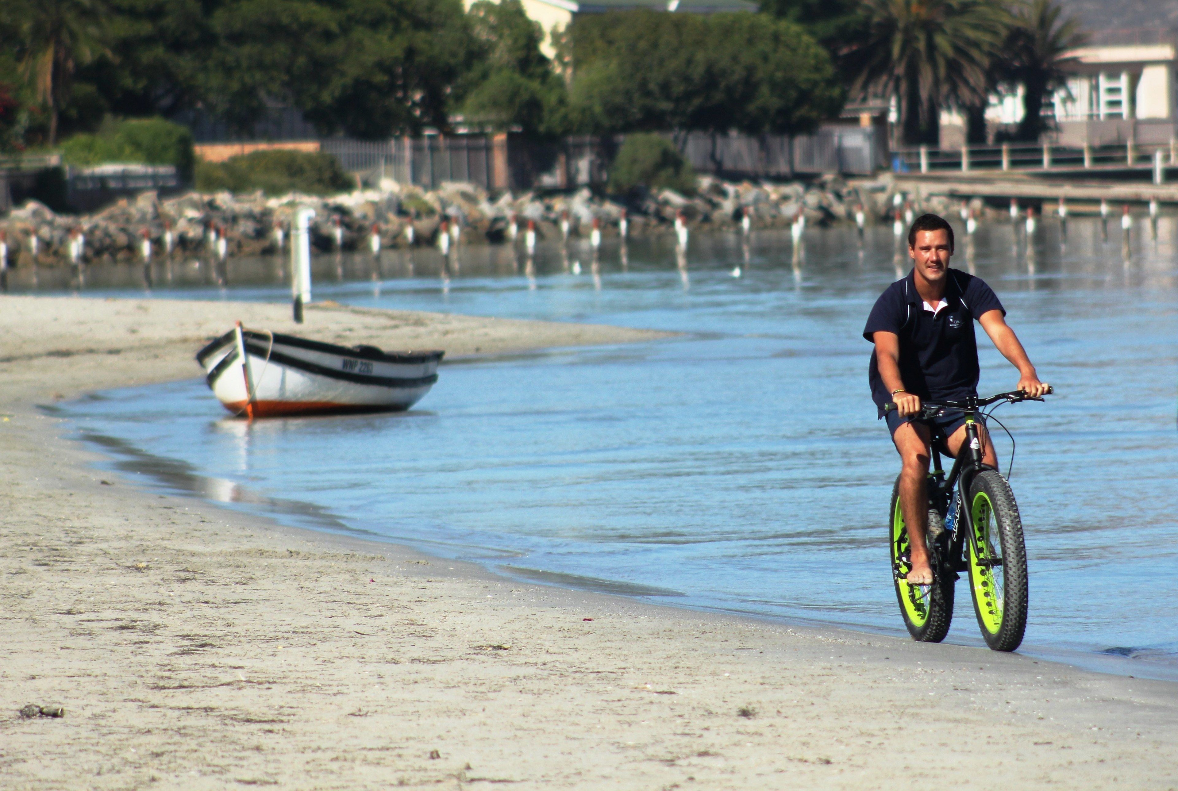A person riding a bicycle on a sandy beach with a boat in the background.