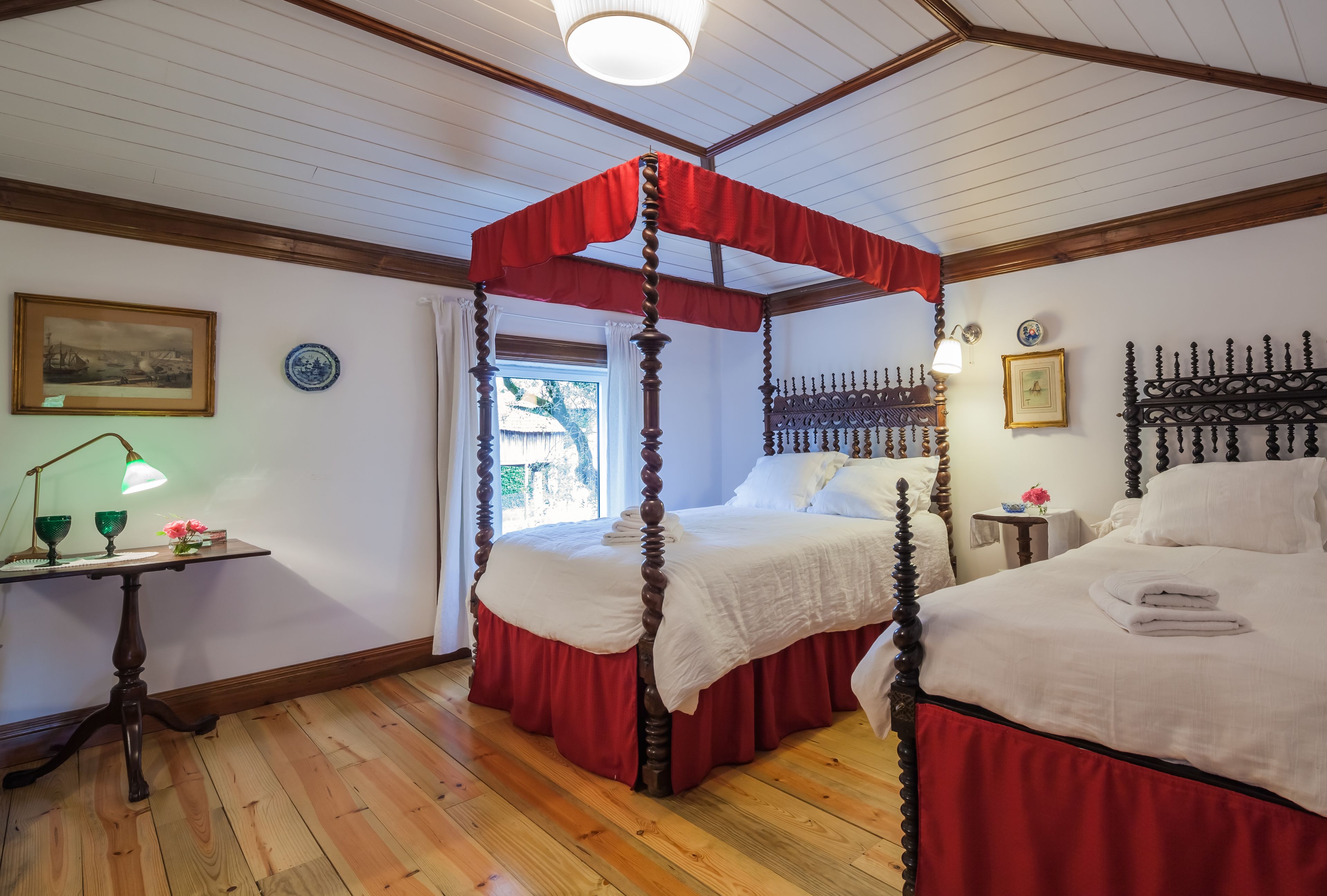 Un lit à baldaquin avec des rideaux rouges, un plancher en bois, une petite table avec une lampe et un tableau encadré au mur.