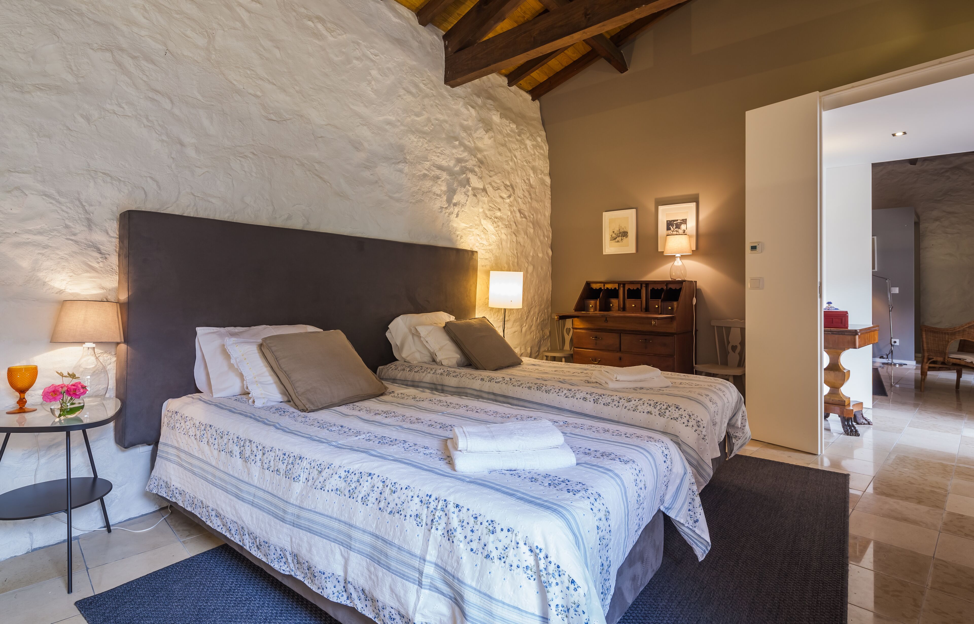 A bedroom with two beds, a nightstand with a lamp, a small table with a vase of flowers, and a wooden cabinet.