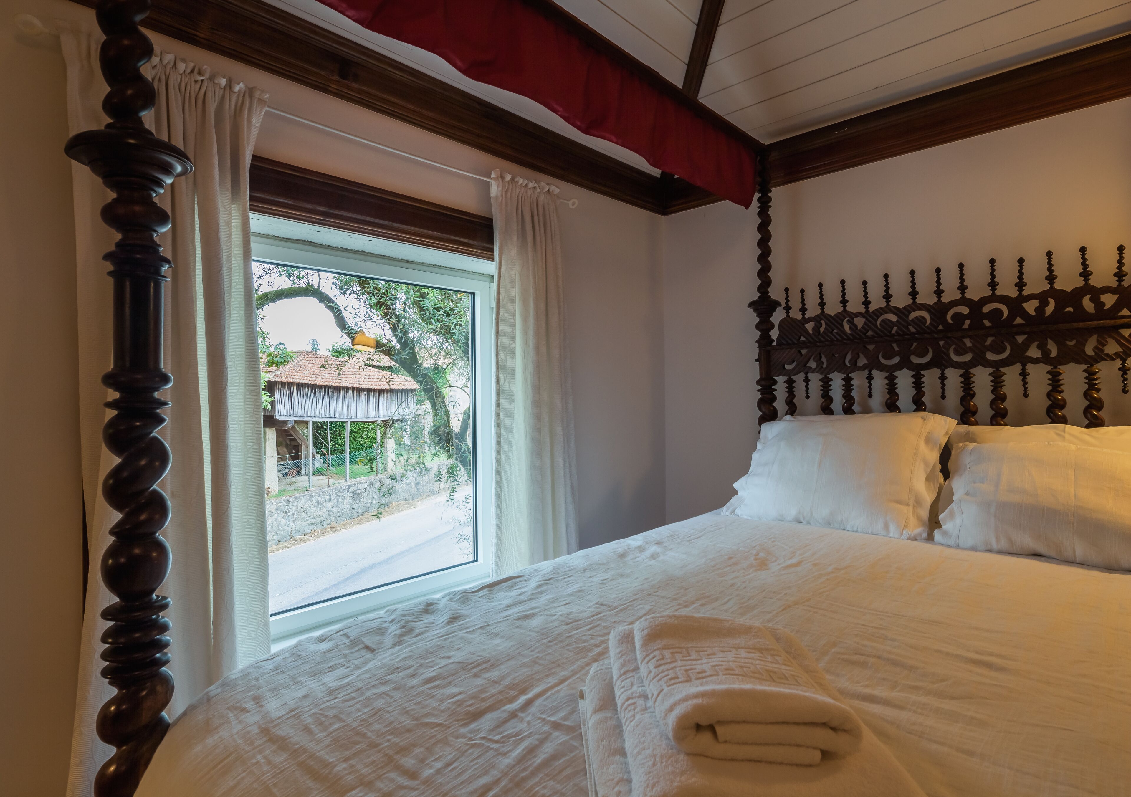 Una cama bien hecha con ropa de cama blanca y una toalla doblada, un cabecero de madera y una ventana con cortinas.