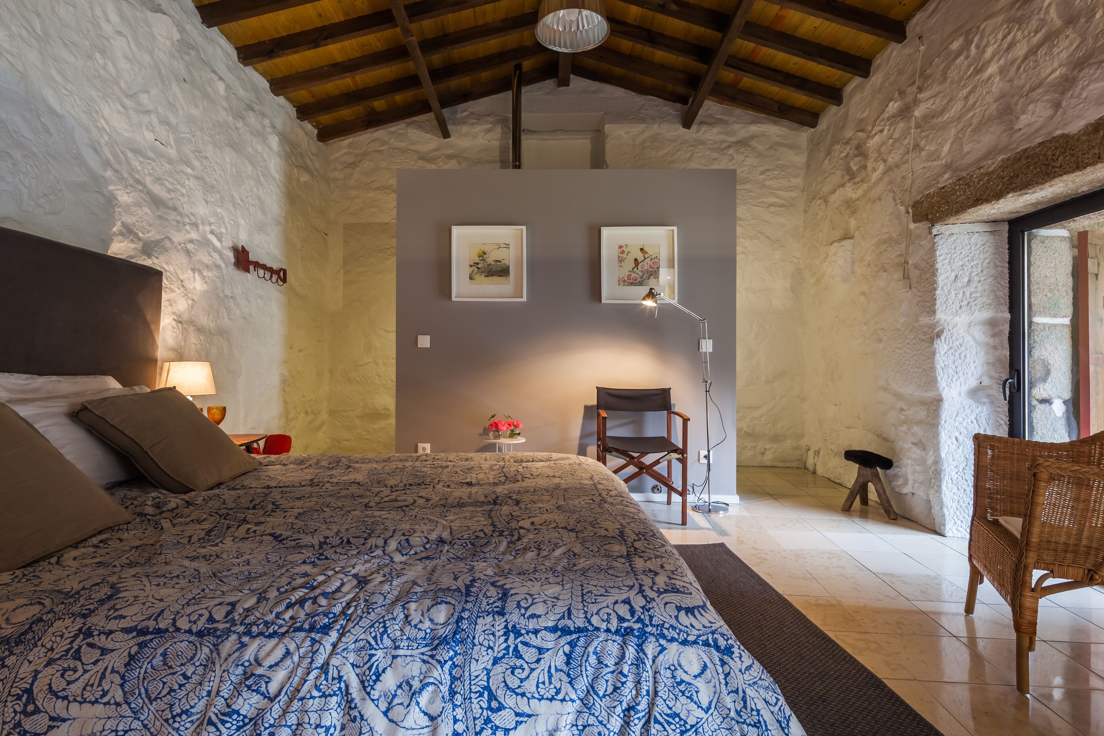 Un dormitorio con una cama grande, techo de madera, una mesita con lámpara y dos cuadros en la pared.