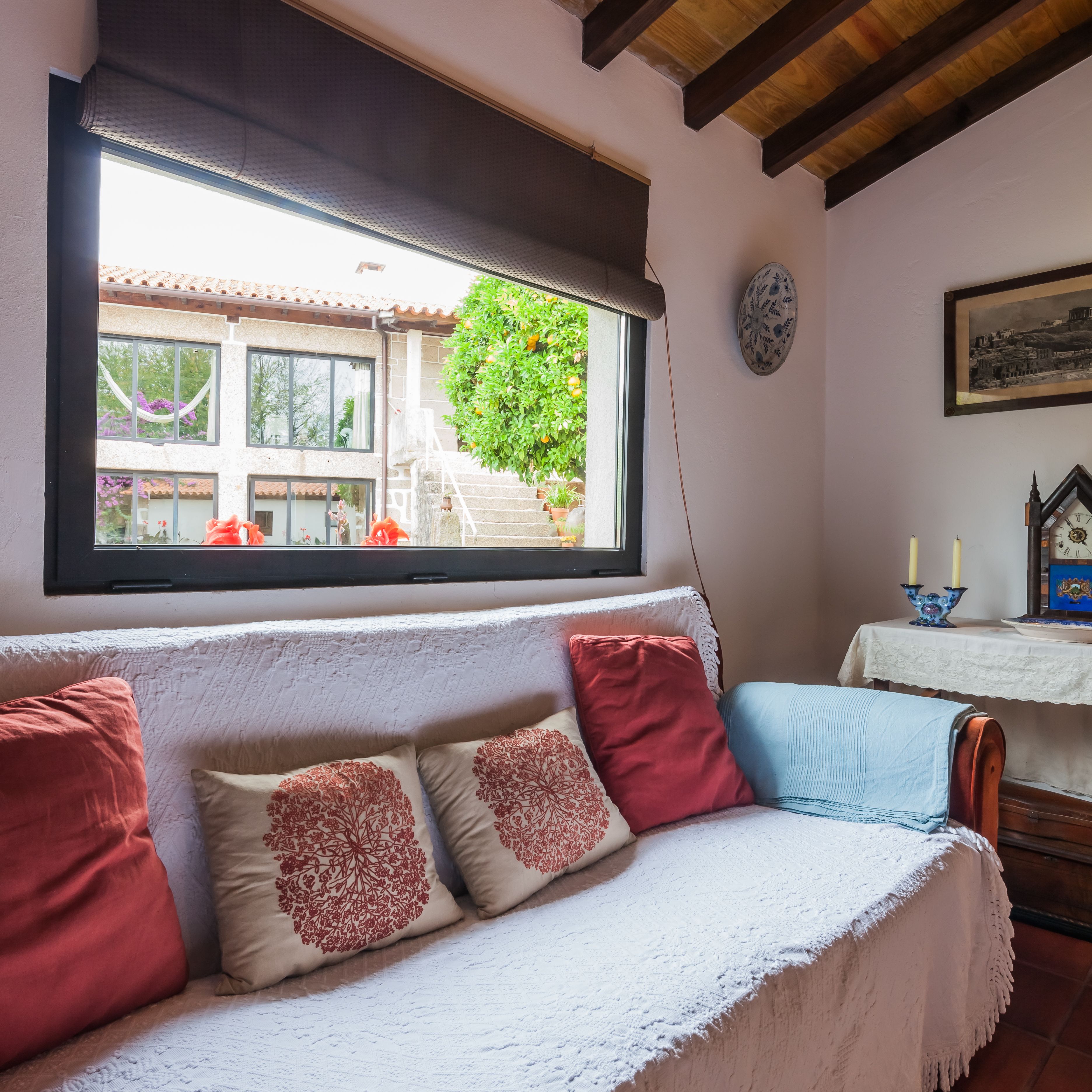 Ein gemütliches Zimmer mit einem weißen Sofa, roten und beigen Kissen, einem Fenster mit Blick auf ein Gebäude und Grünflächen, einer Uhr an der Wand und einem Tisch mit Kerzen.