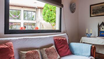 Ein gemütliches Zimmer mit einem weißen Sofa, roten und beigen Kissen, einem Fenster mit Blick auf ein Gebäude und Grünflächen, einer Uhr an der Wand und einem Tisch mit Kerzen.