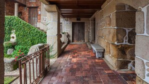 Un chemin pavé de briques, partiellement abrité, mène à une porte en bois, le tout étant entouré de murs en pierre et d’une clôture en métal.