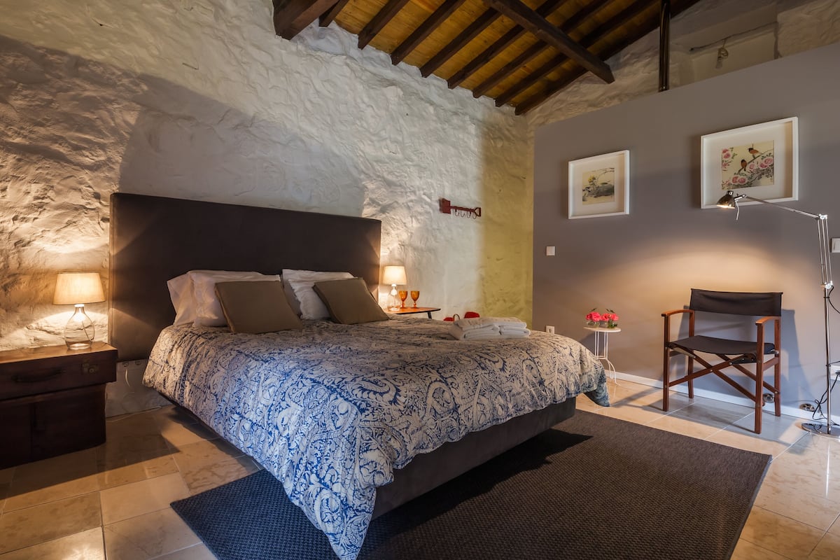 Un dormitorio con una cama grande, una mesita de noche, una lámpara, una silla y cuadros en la pared.