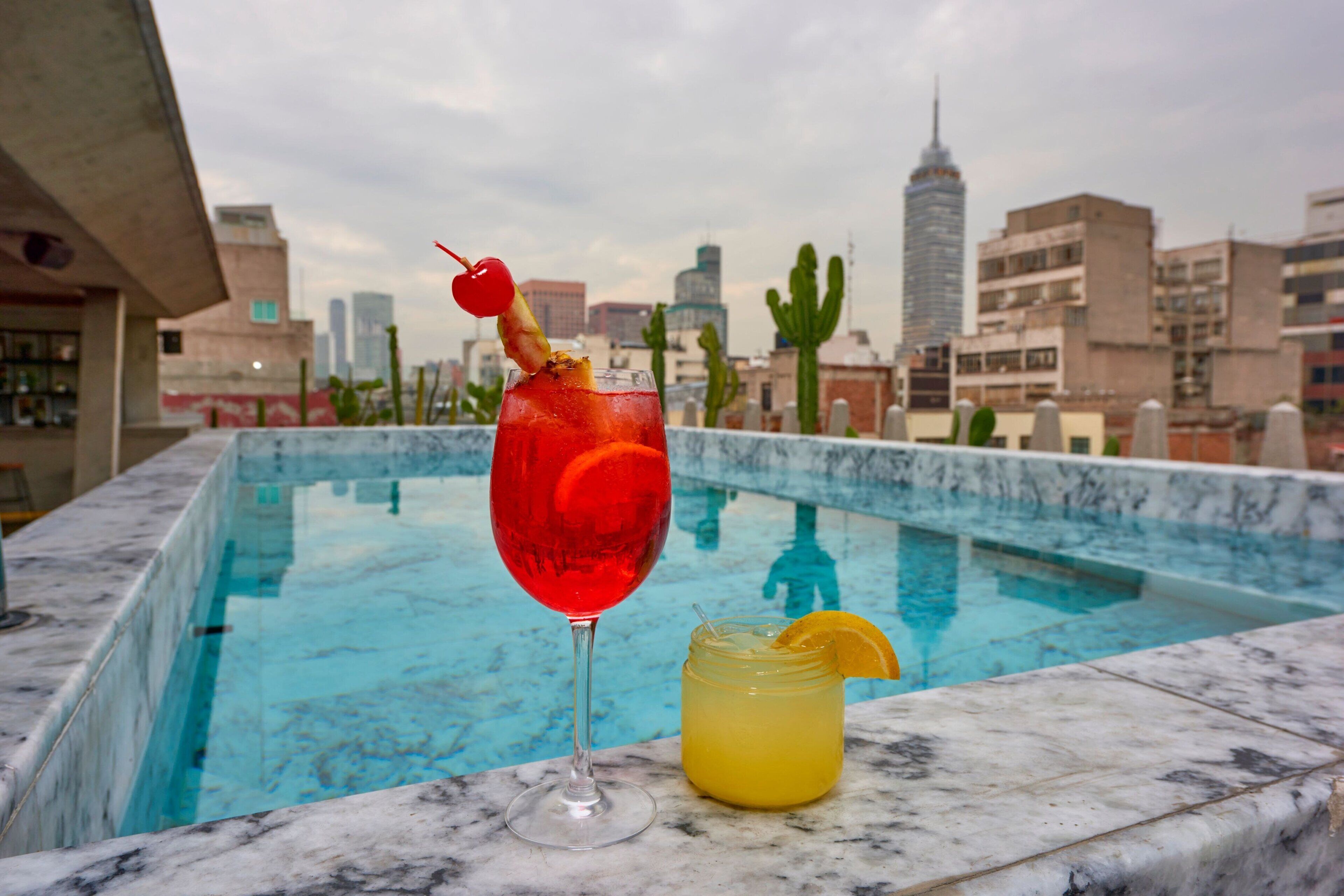 A poolside setting with a drink in a glass and a mason jar, overlooking a cityscape with buildings and cacti.