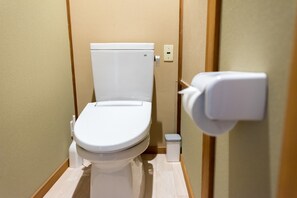 Baño de hotel estándar con inodoro blanco, un pequeño cubo de basura y un accionador de descarga en la pared.