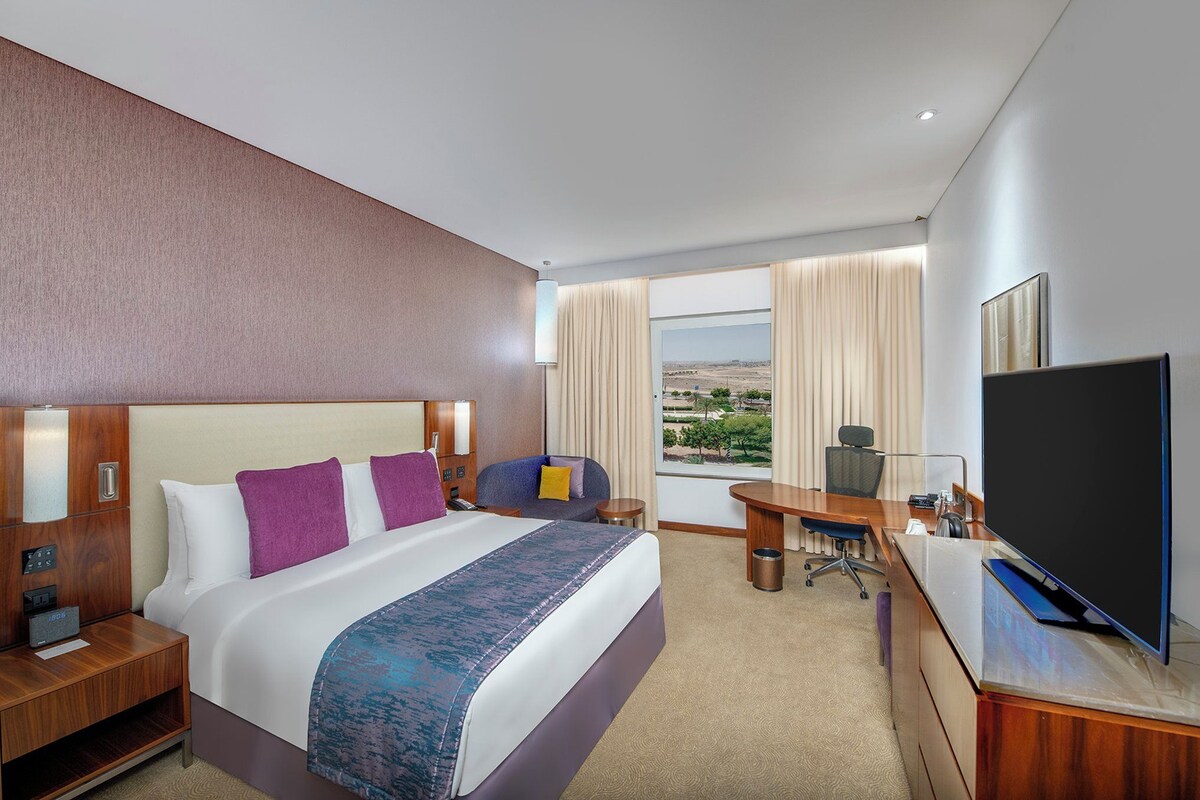 A hotel room with a large bed, a desk with a computer, a TV, and a view of the desert.