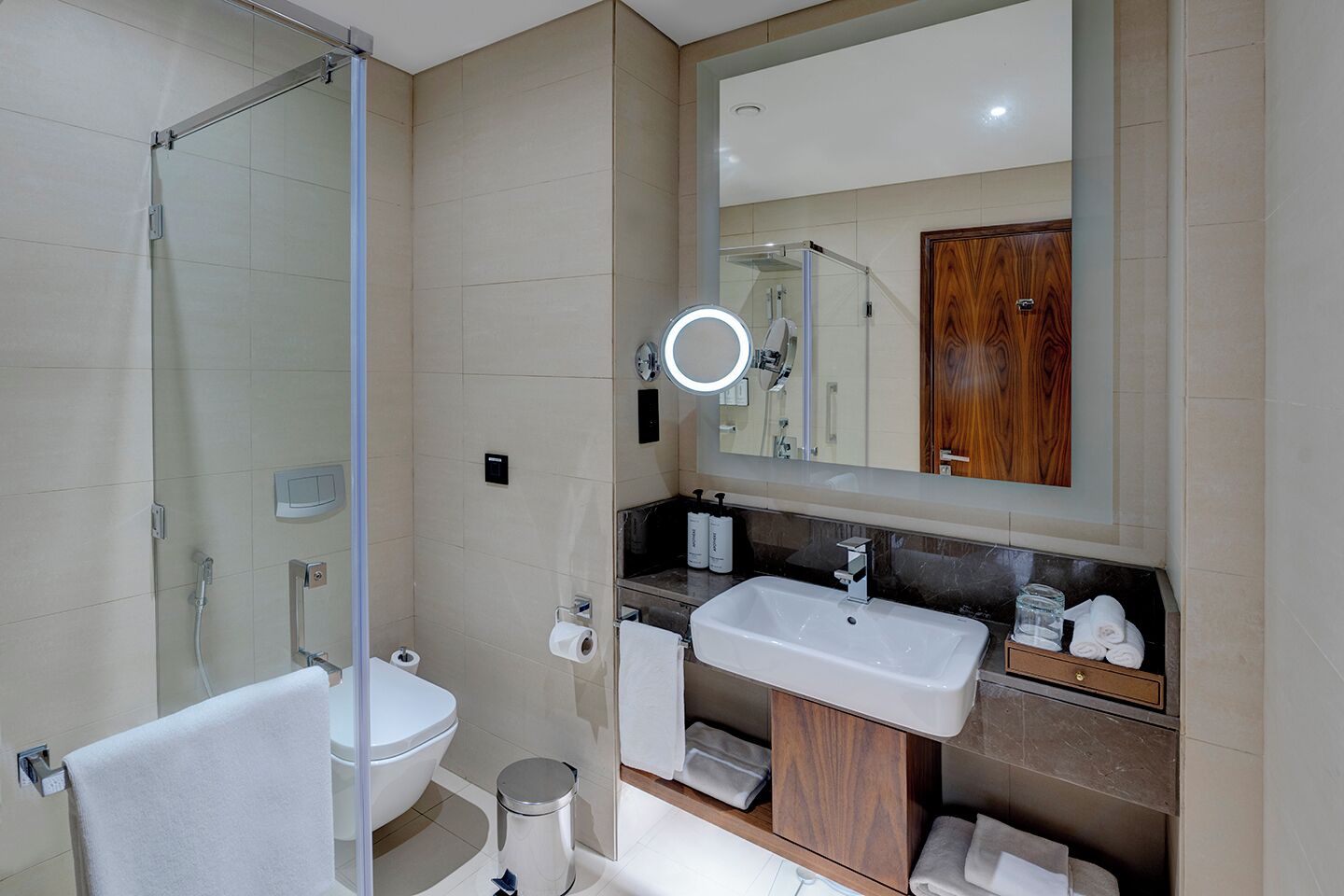 Ein modernes Badezimmer mit Duschkabine aus Glas, Toilette in Weiß, Waschbecken mit Holzschrank und Spiegel mit runder Beleuchtung.
