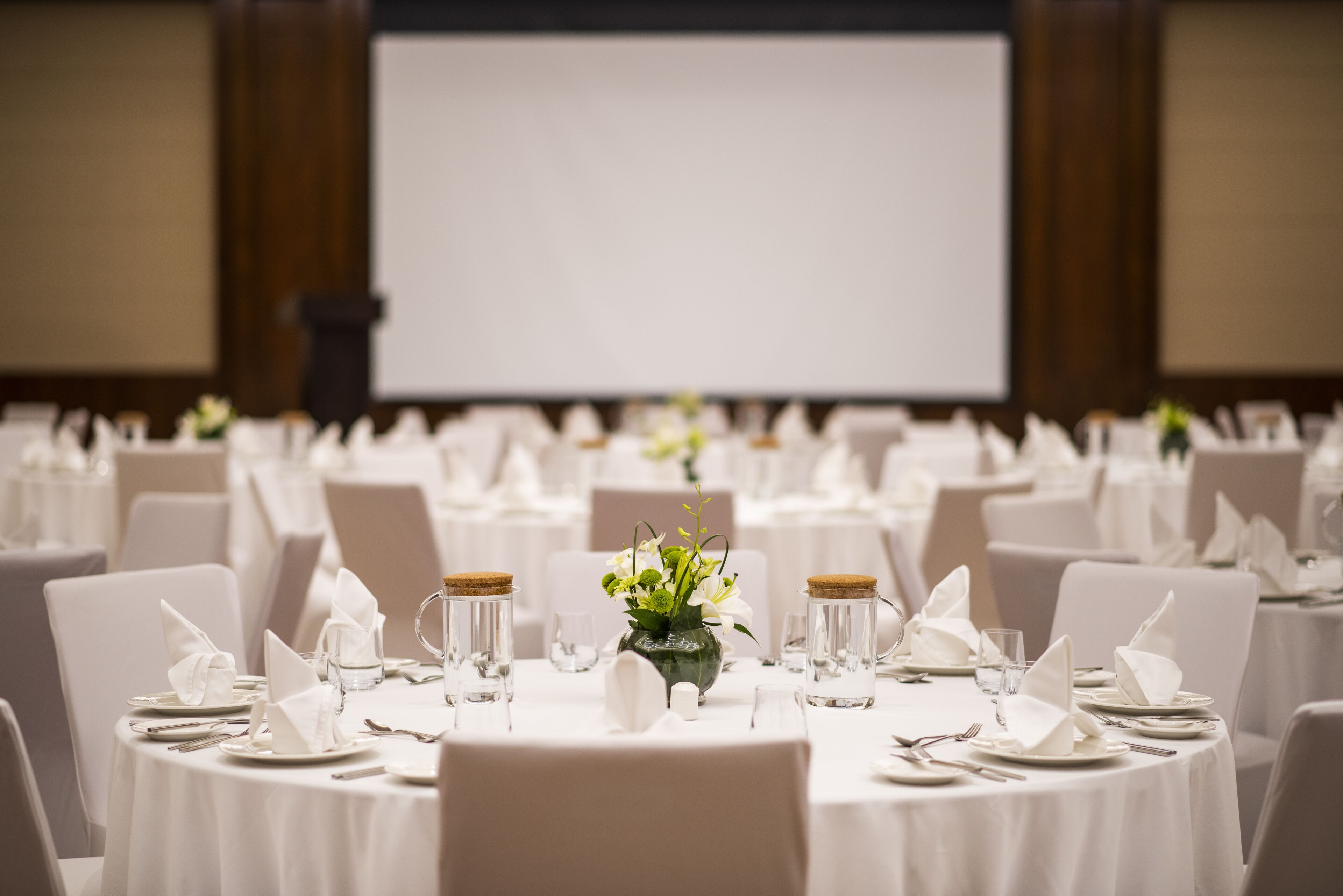 A banquet hall with round tables set for a formal event, featuring white tablecloths, folded napkins, and clear glassware.