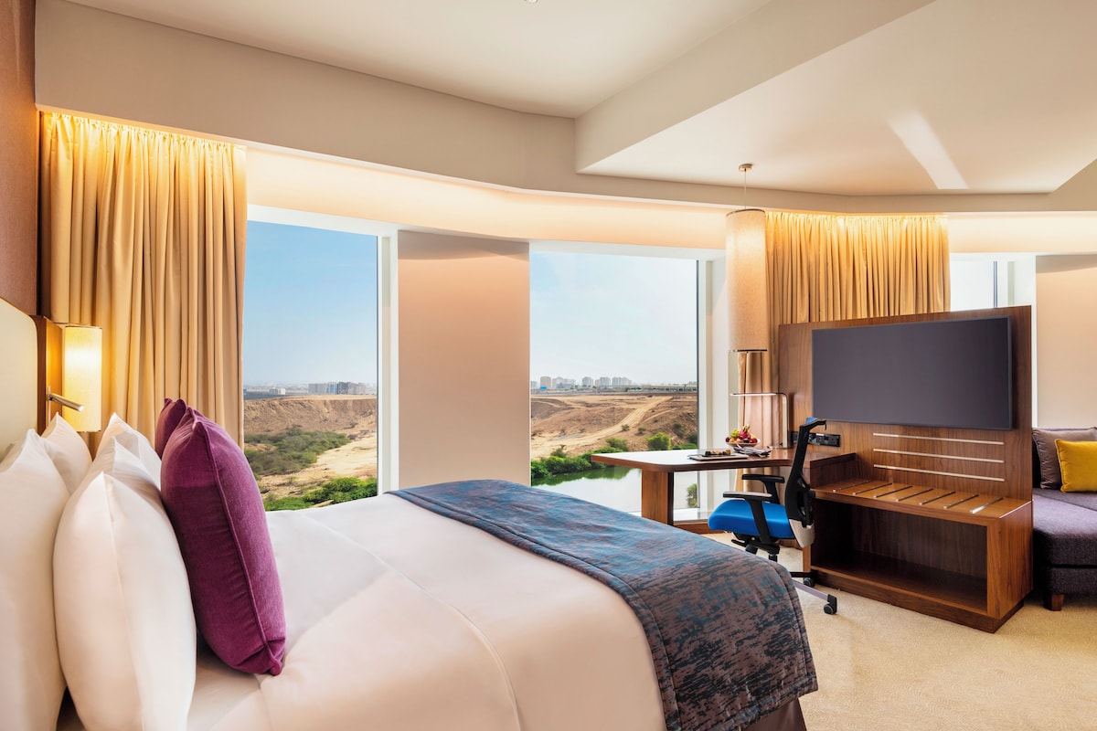 A hotel room with a bed, a desk with a flat-screen TV, a chair, and a view of a landscape.