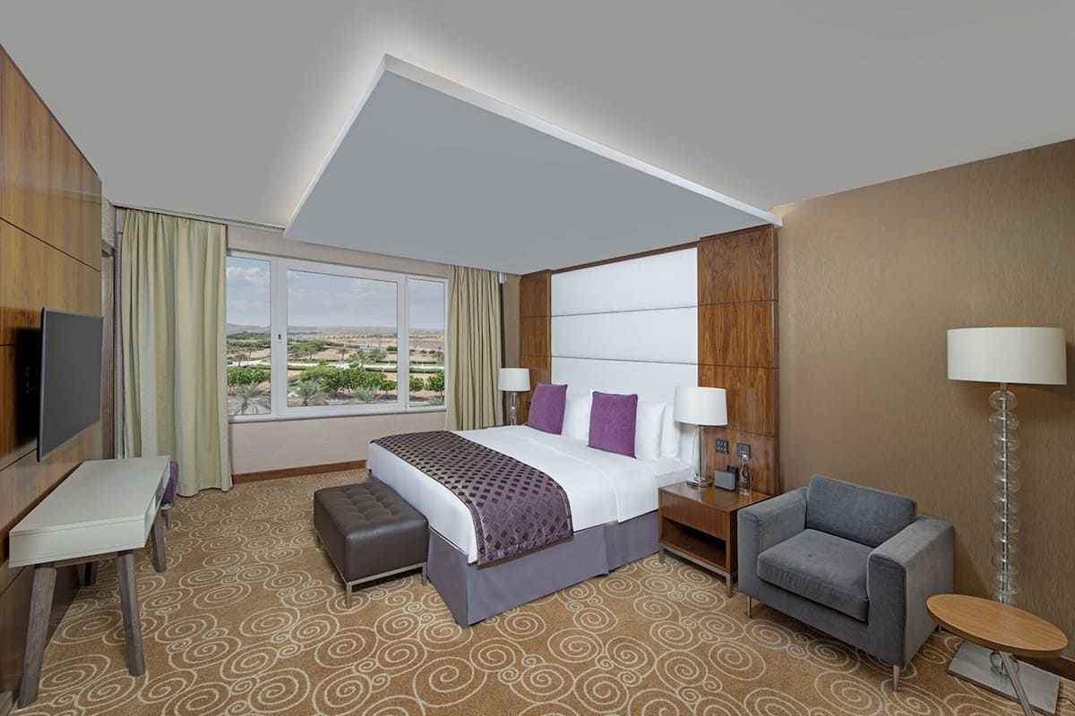 A hotel room with a large bed, a desk, a chair, a small table, and a TV.