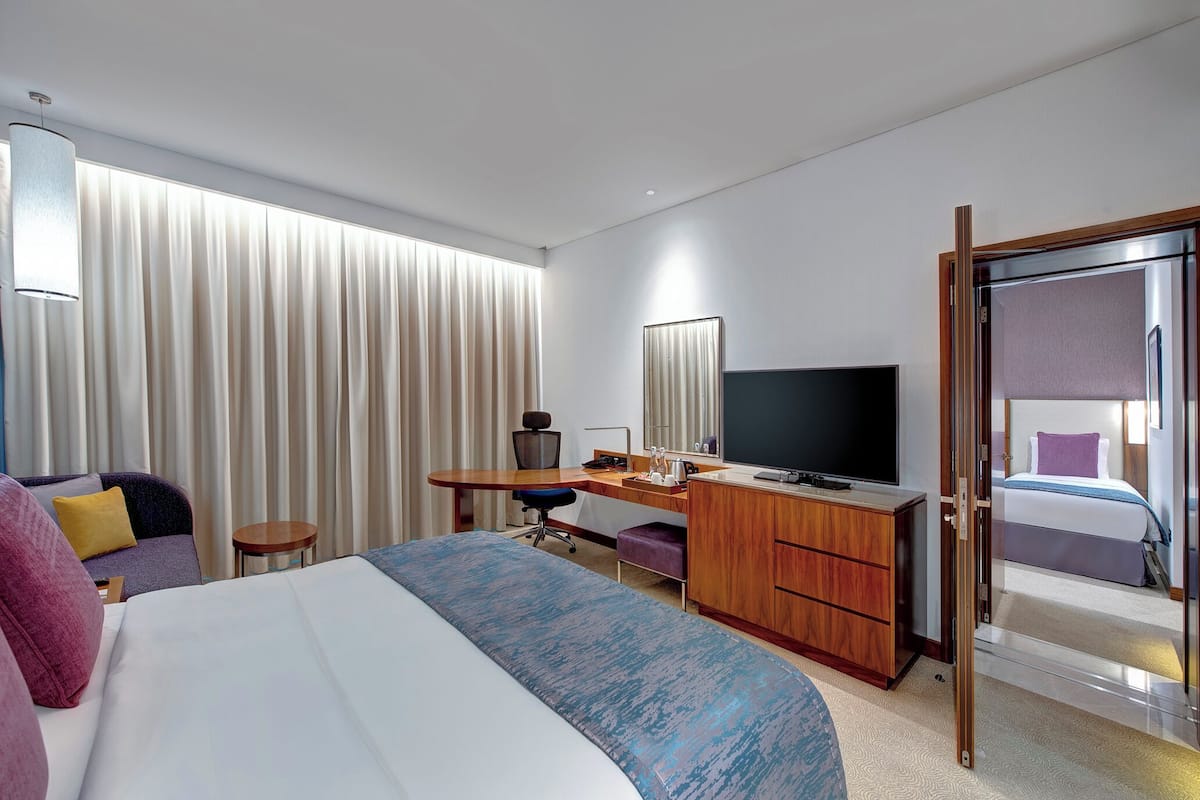 A hotel room with a bed, a desk with a computer, a chair, a TV, and a large window with curtains.