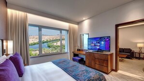 Habitación de hotel con una cama grande, un escritorio con televisor y vistas a un río y al paisaje urbano.