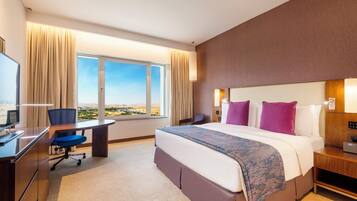 A hotel room with a large bed, a desk, a chair, a TV, and a view of the landscape.