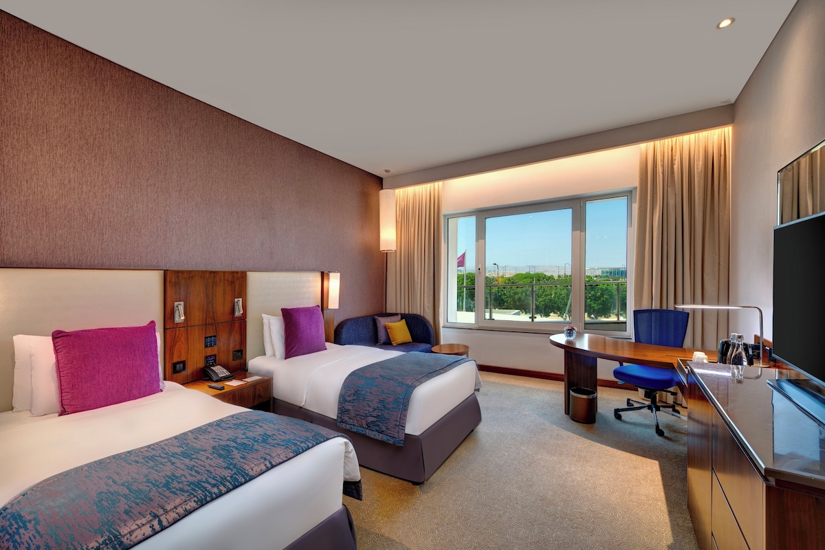A hotel room with two beds, a desk, a chair, a TV, and a large window with a view.