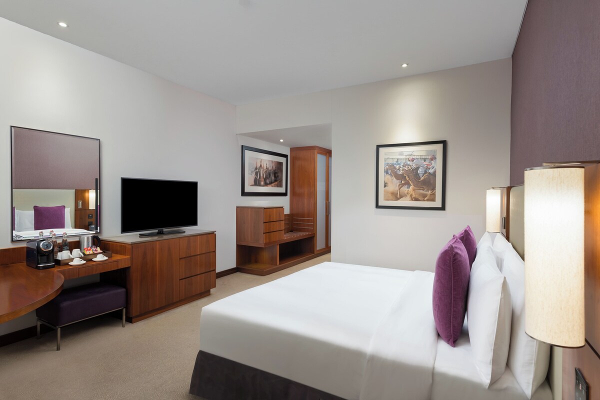 A hotel room with a bed, a desk with a television, a wardrobe, and a large framed picture on the wall.