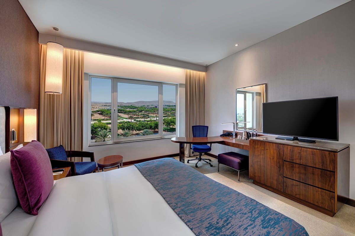 A hotel room with a large bed, a desk with a flat-screen TV, a chair, a small table, and a view of the outdoors.