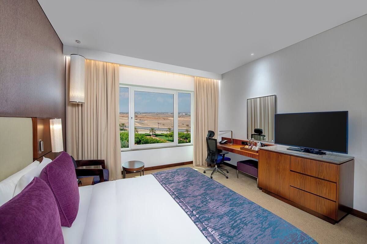 A hotel room with a large bed, a desk with a computer, a TV, and a window with a desert view.
