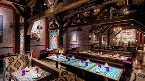 Un restaurante con vigas de madera, paredes con diseños y mesas dispuestas con servilletas y platos morados.
