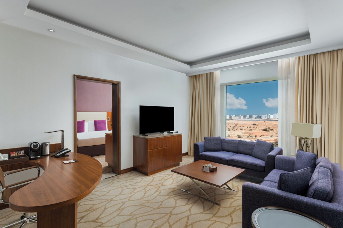 A hotel room with a bed, a TV, a sofa, a coffee table, a desk with a chair, and a view of a desert landscape.