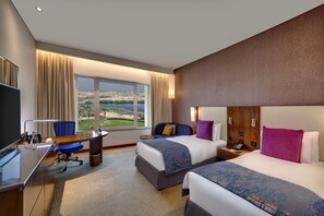 Relax in a spacious room with a large bed, a smaller bed, and a desk area, offering a view of a serene landscaped area with a body of water.
