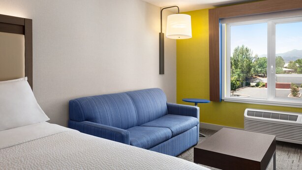 Habitación de hotel con cama, sofá azul, una mesita, ventana con vistas a zonas verdes y montañas, y una lámpara fijada a la pared.
