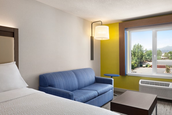 A hotel room with a bed, a blue sofa, a small table, a window with a view of greenery and mountains, and a wall-mounted lamp.