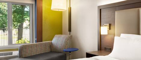 A hotel room with a bed, a chair, a small table, a lamp, and a window with a view of trees.