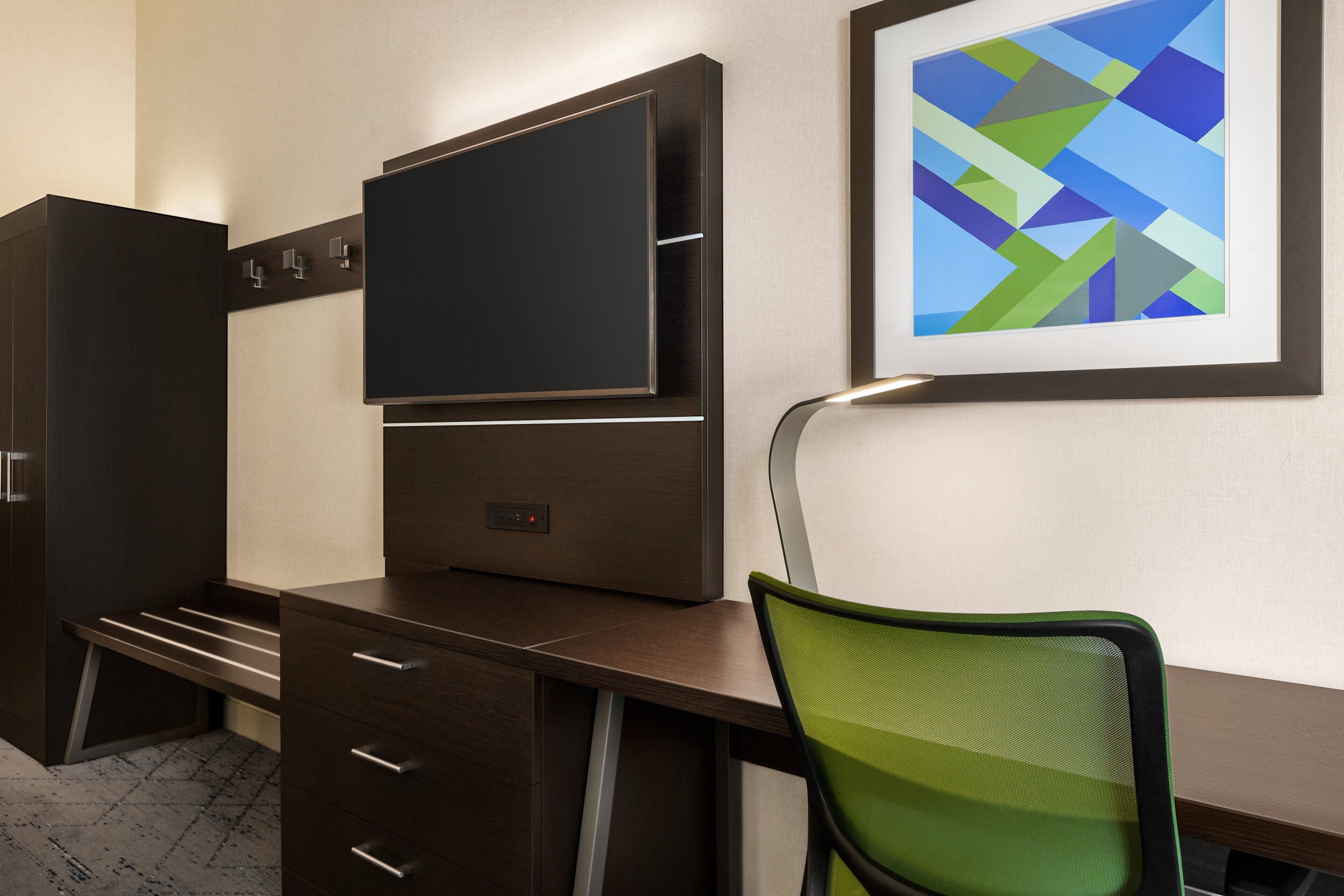 A modern office desk with a flat-screen TV, a framed abstract painting, and a green mesh chair.