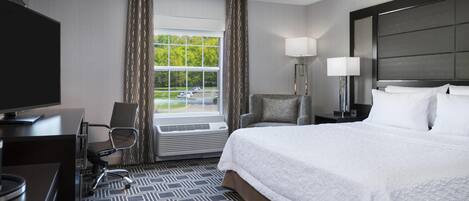 A hotel room with a bed, desk, chair, TV, window, and lamp.