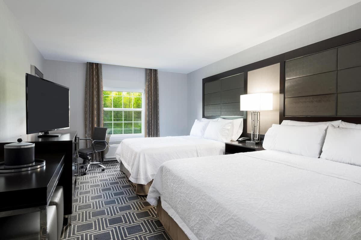Quarto de hotel com duas camas, uma escrivaninha com televisão, uma cadeira, uma janela com cortinas e cabeceira com luminária.