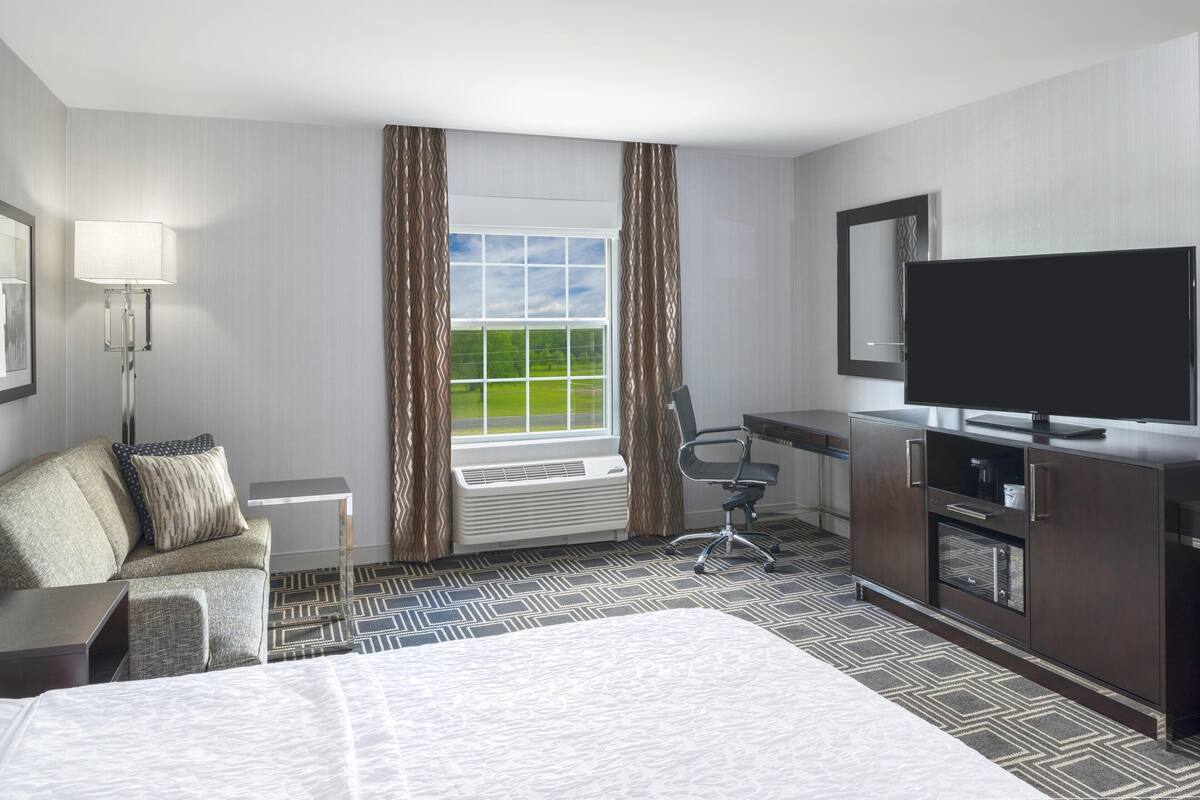 Quarto de hotel com cama, escrivaninha, cadeira, televisão e janela com cortinas.