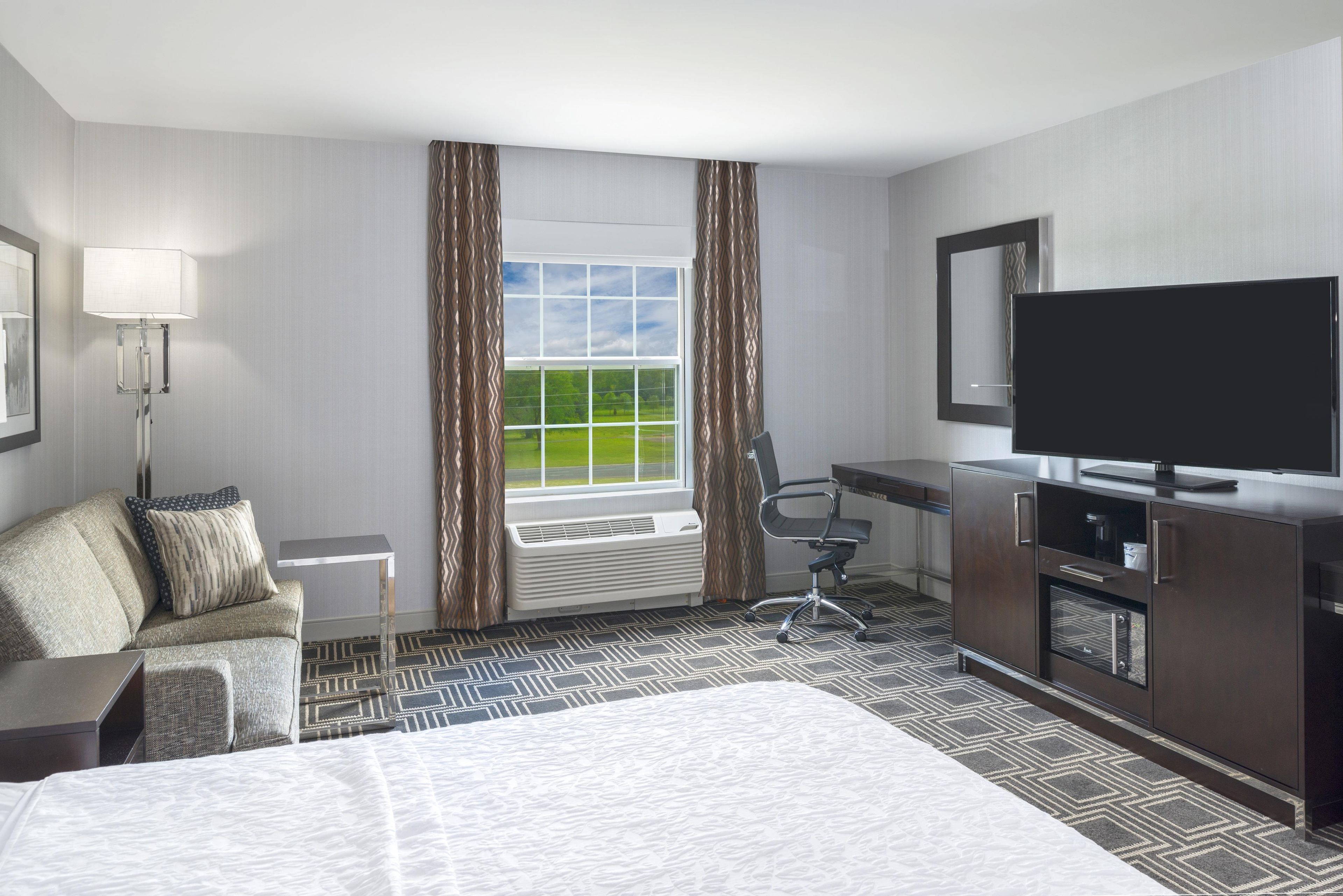 Ein Hotelzimmer mit einem Bett, einem Schreibtisch, einem Sessel, einem Fernseher und einem Fenster mit Vorhängen.