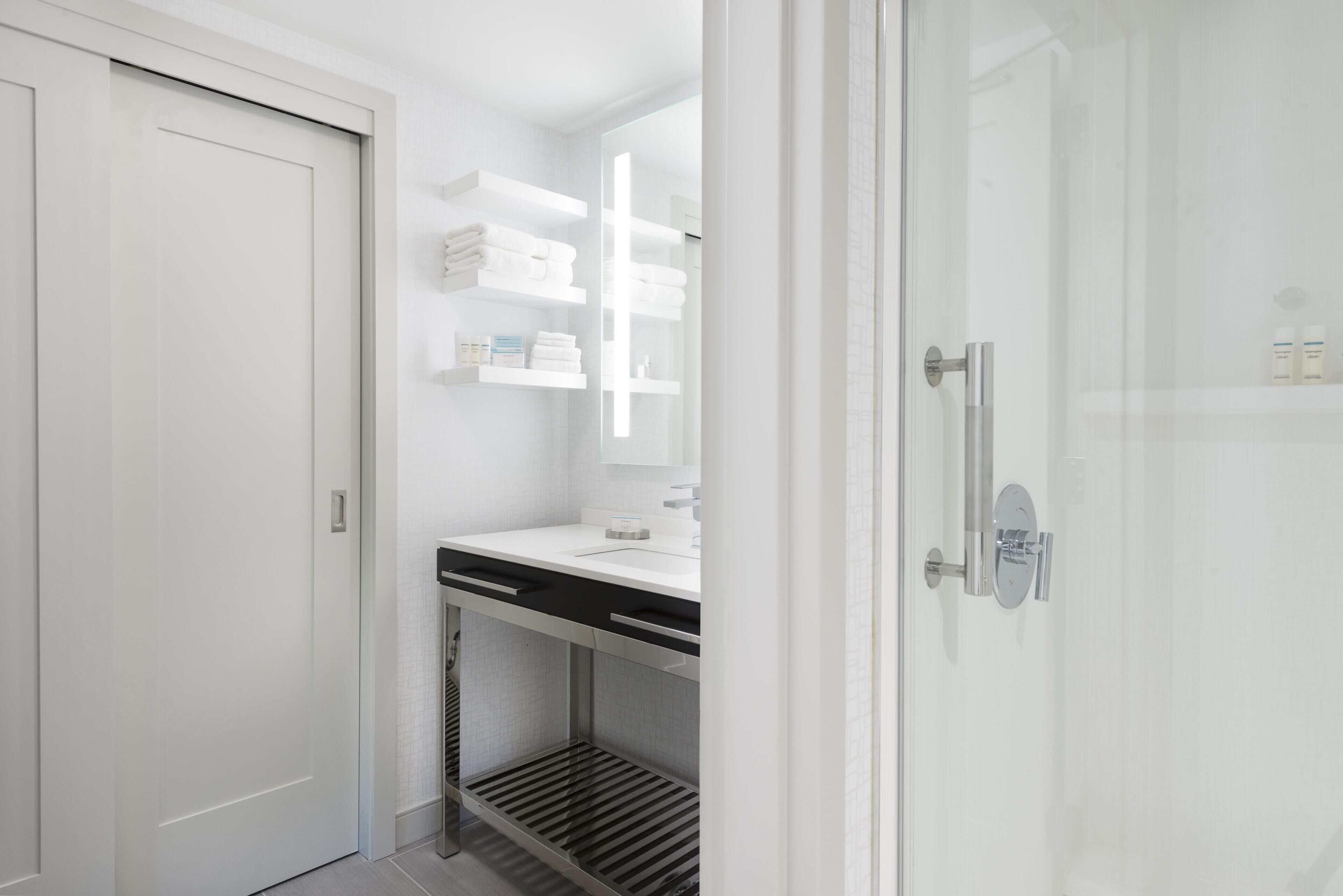 A modern bathroom with a white door, a glass shower enclosure, a white countertop, and a shelf with towels.