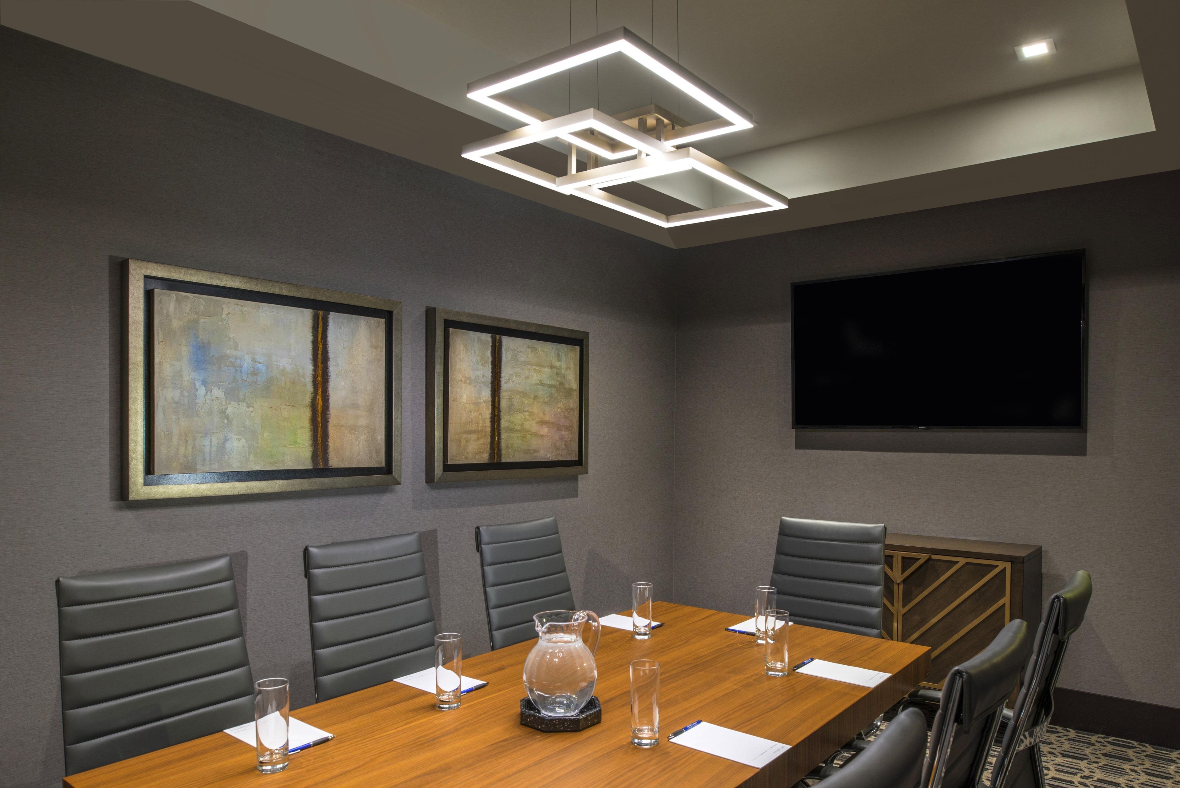 A conference room with a long table, six chairs, two framed abstract paintings, a flat-screen TV, and a modern ceiling light fixture.
