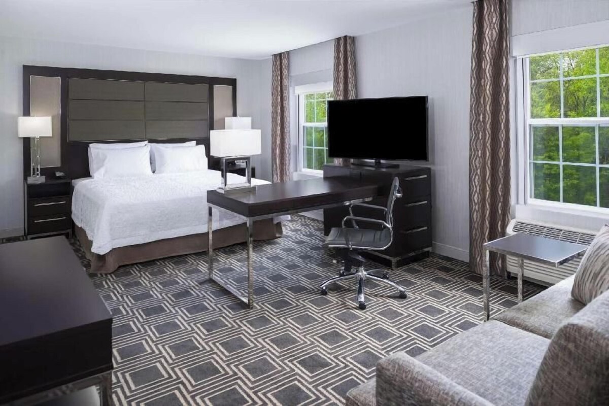 A hotel room with a bed, desk, chair, TV, and a window with curtains.