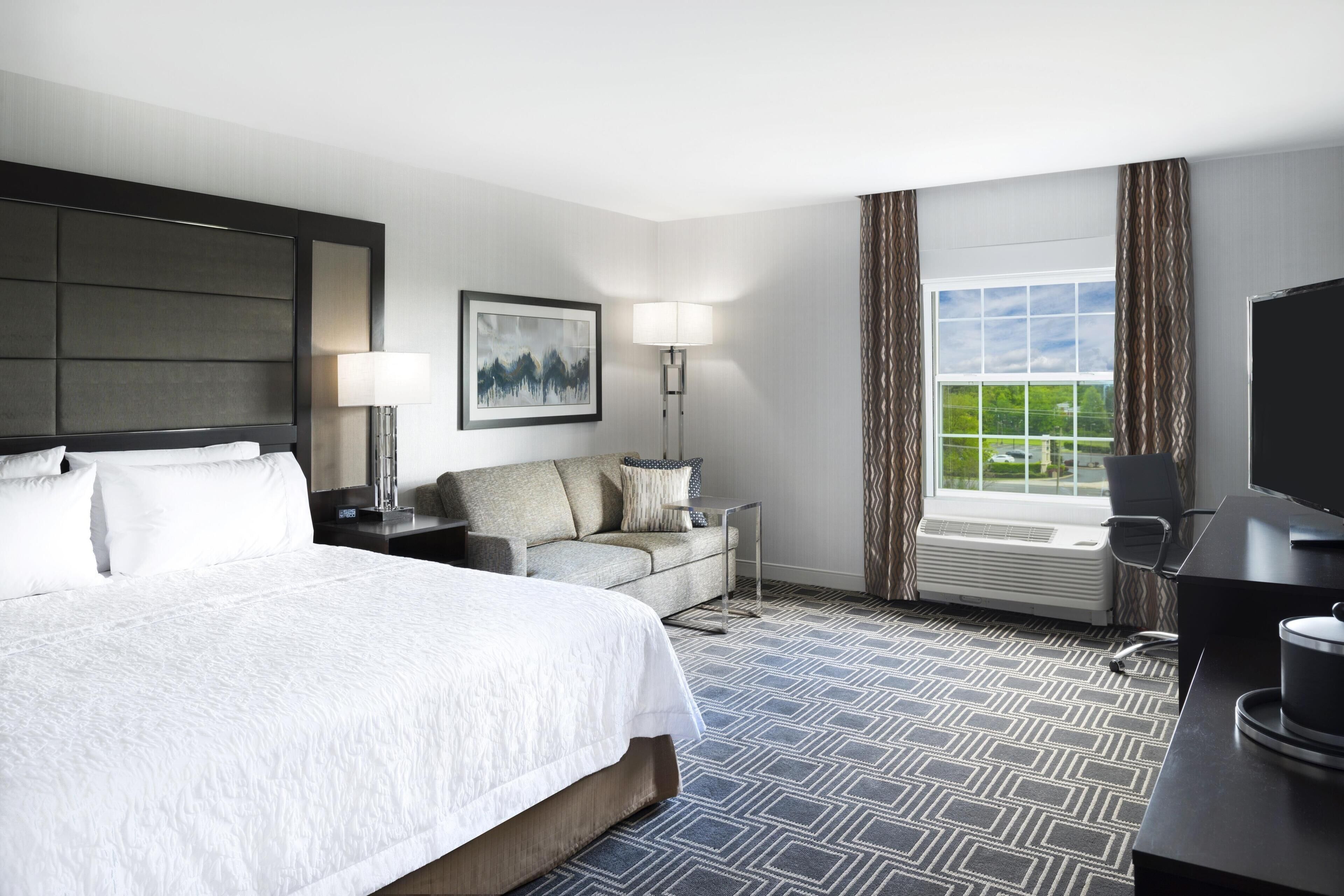 A hotel room with a large bed, a sofa, a desk, a lamp, a painting, a window with curtains, and a view of greenery.
