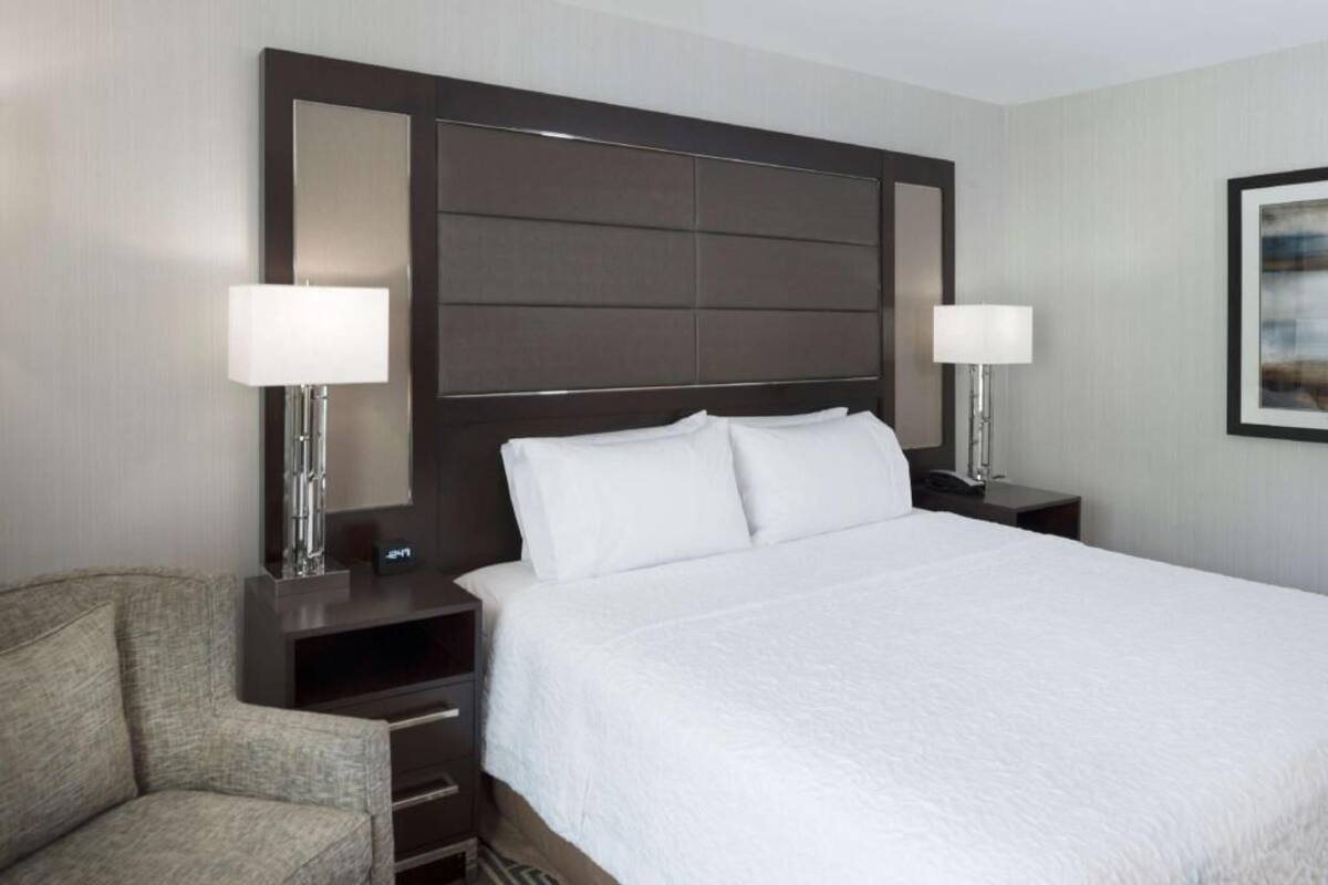 A hotel room with a large bed, two bedside tables with lamps, a chair, and a large mirror on the headboard.