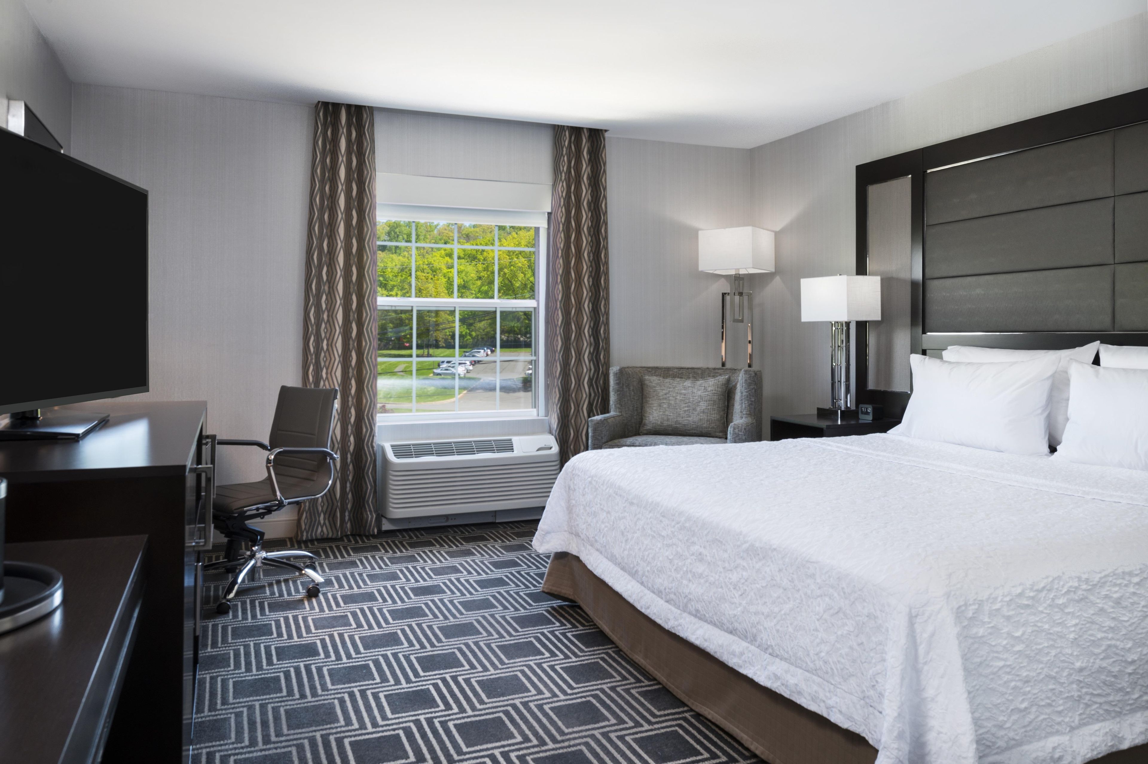 A hotel room with a bed, a desk, a chair, a TV, a window with curtains, and a view of the outdoors.