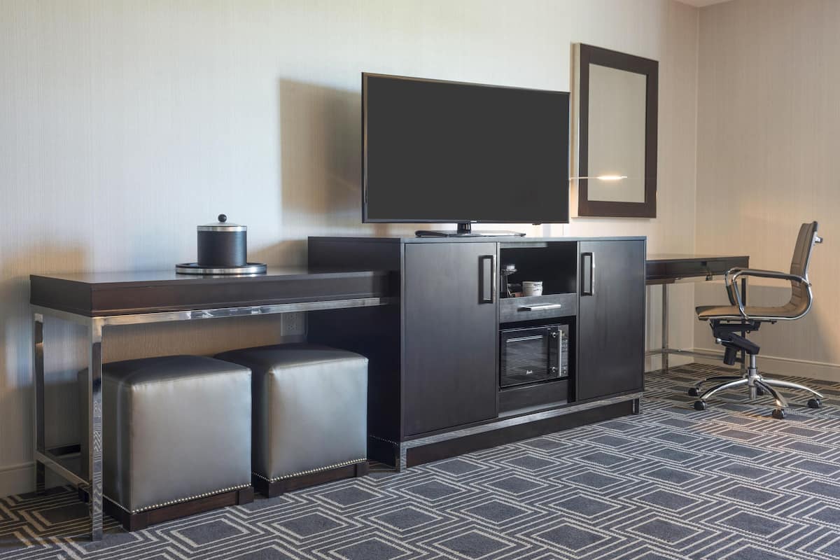 Quarto de hotel moderno com TV de tela plana, forno embutido, cafeteira, mini-geladeira, escrivaninha com computador e uma cadeira.