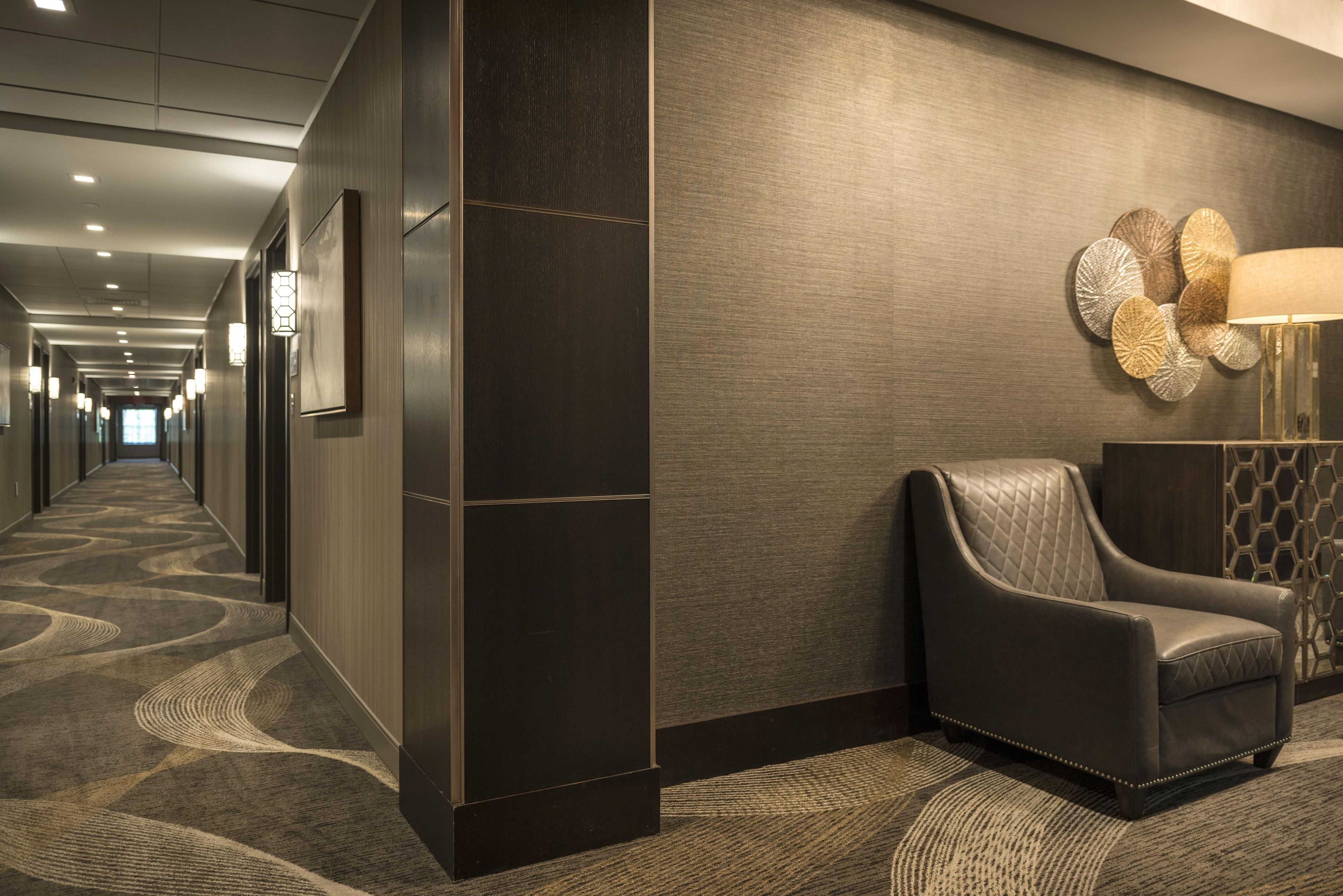 Corredor de hotel com carpete estampado, paredes escuras e um espaço de estar com poltrona de couro e luminária decorativa.