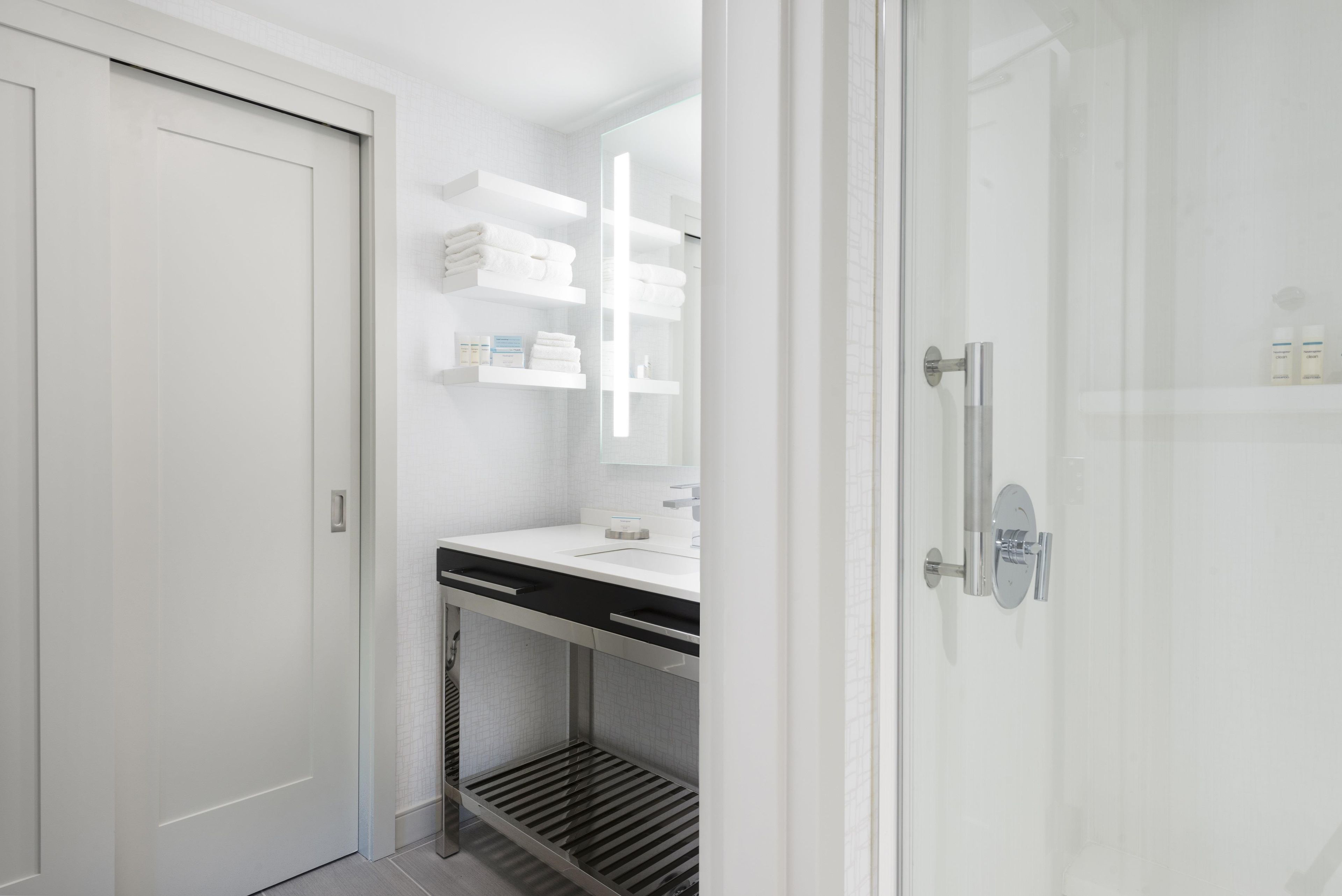 Ein modernes Badezimmer mit einer weißen Tür, einer Duschkabine aus Glas, einem weißen Waschbecken mit Spiegel und einem Handtuchhalter mit gefalteten Handtüchern.