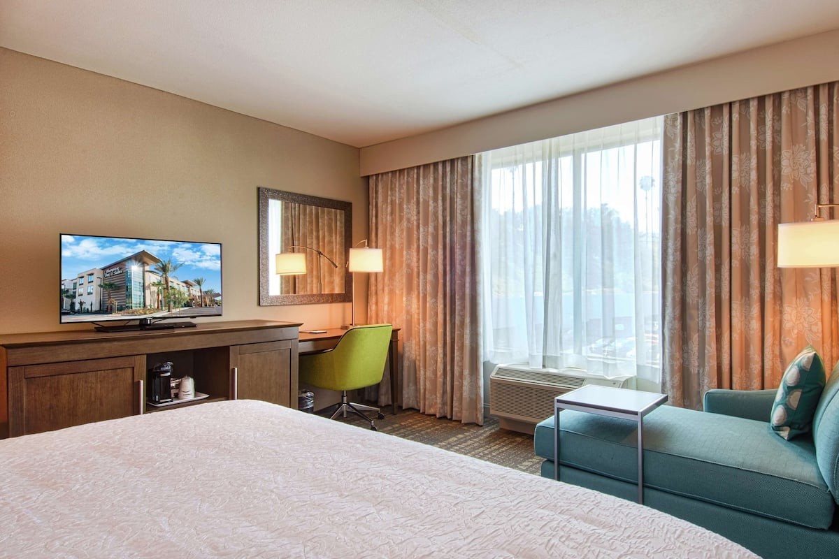 Una camera d'albergo con un letto, una scrivania, una sedia, una televisione, uno specchio e una finestra con tende.