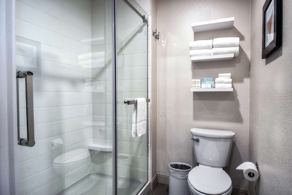 A modern bathroom with a glass shower enclosure, a toilet, and a shelf with towels and toiletries.