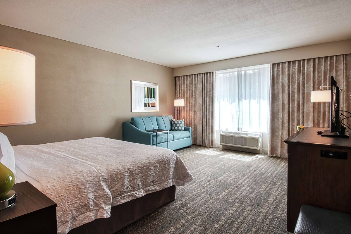 Una camera d'albergo con un letto, un divano, una scrivania, una lampada e un'ampia finestra con tende.