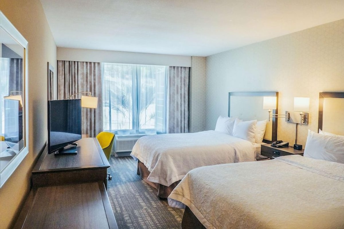A hotel room with two beds, a desk with a computer, a chair, and a window with curtains.