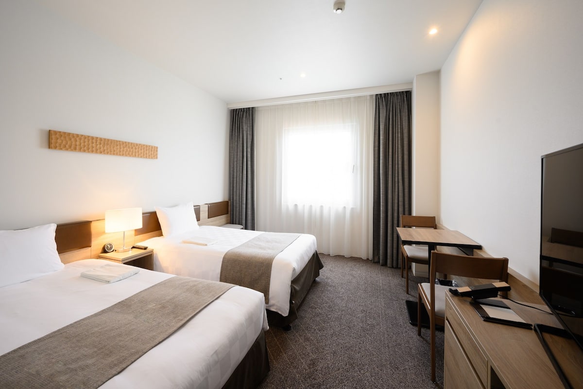 A hotel room with two beds, a desk, a television, and a large window with curtains.
