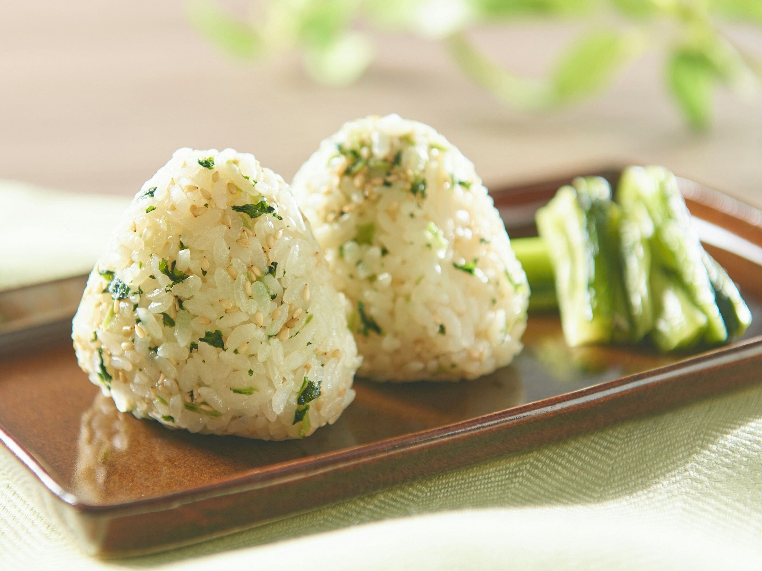 Two rice balls with green herbs and sesame seeds on a rectangular plate, accompanied by a side of green vegetables.