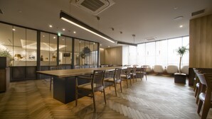Een moderne vergaderruimte met een lange tafel en stoelen, houten visgraatvloer en grote ramen met zonwering.